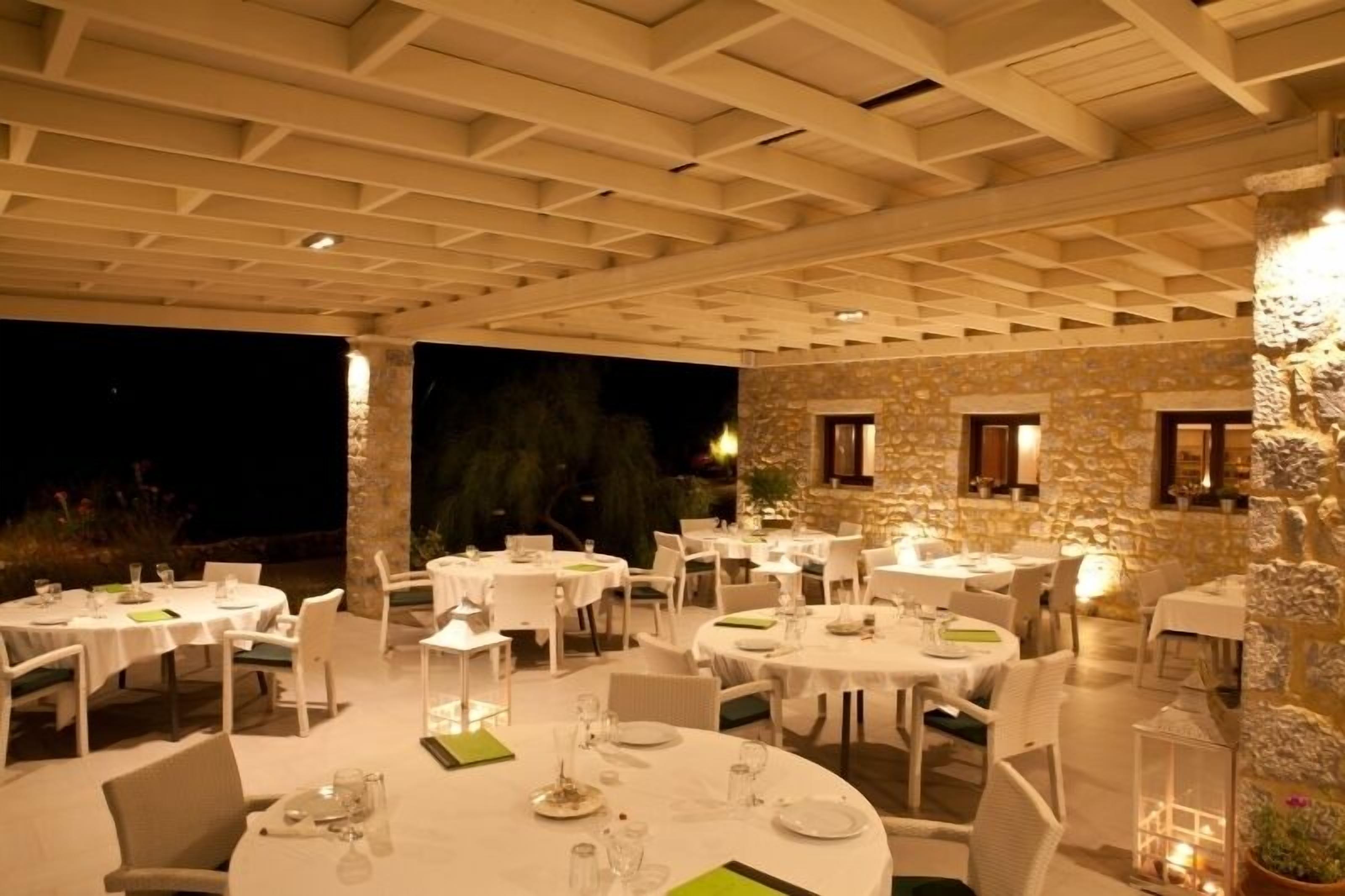 An outdoor dining area with round tables and chairs, stone walls, and warm lighting.