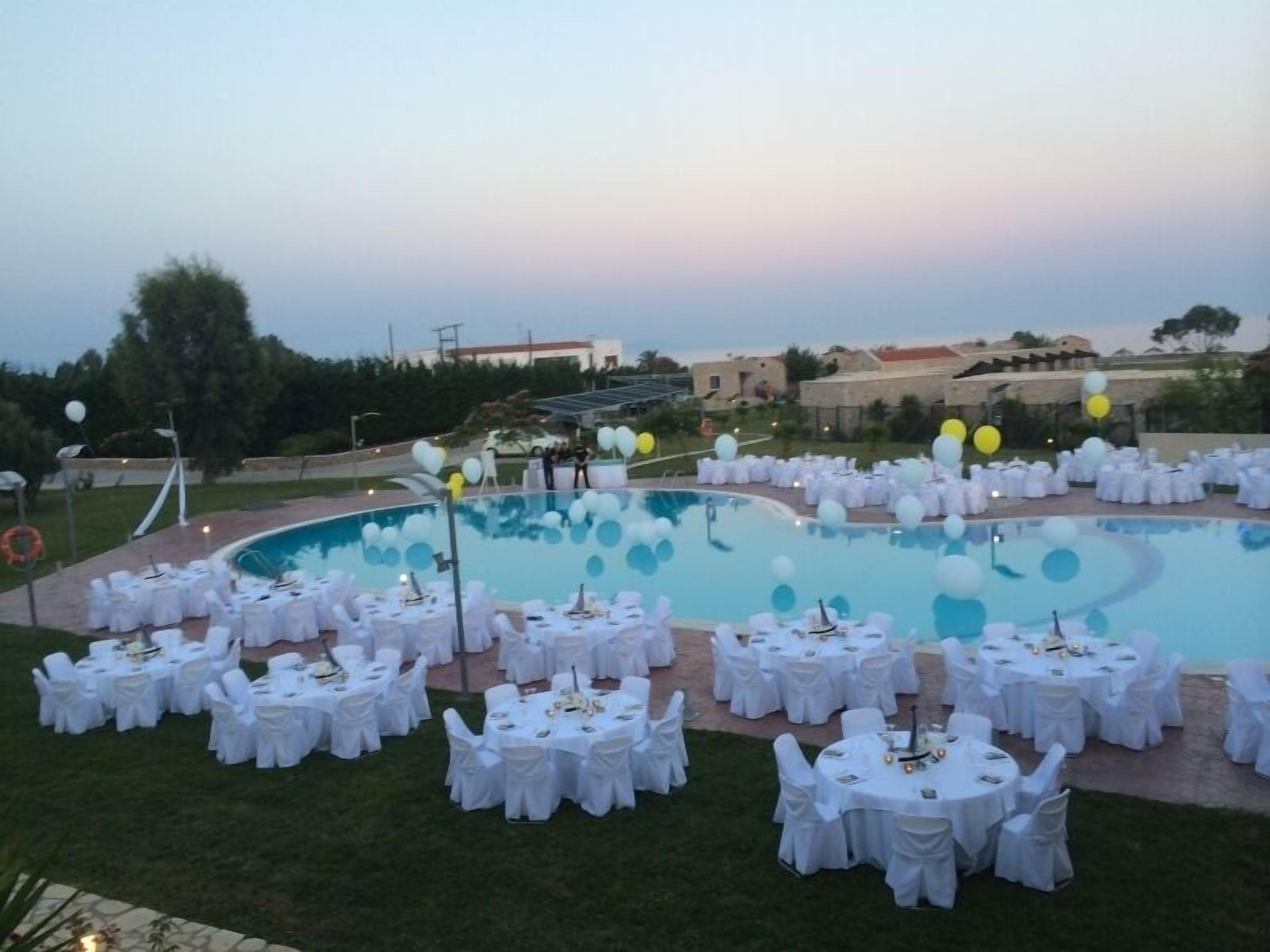 Un área de piscina al aire libre con mesas y sillas redondas, rodeada de manteles blancos y sillas, preparada para un evento.