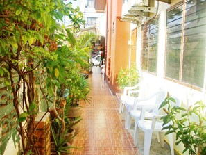 A narrow brick pathway flanked by potted plants and white plastic chairs, leading to a building with large windows. - JJ Guesthouse (Karon)