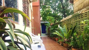 A covered outdoor seating area with white plastic chairs and tables, surrounded by potted plants and a brick wall. - JJ Guesthouse (Karon)