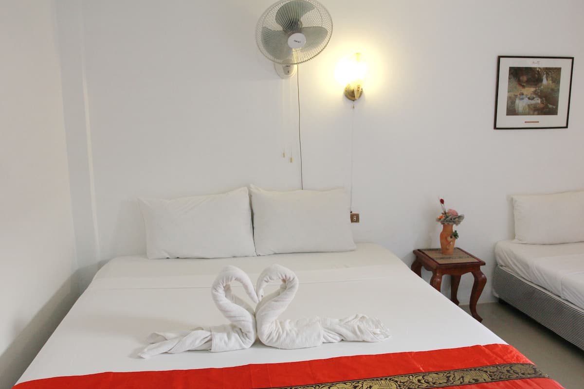 A hotel room with a bed, a wall-mounted fan, a bedside table with a vase, and a framed picture on the wall.