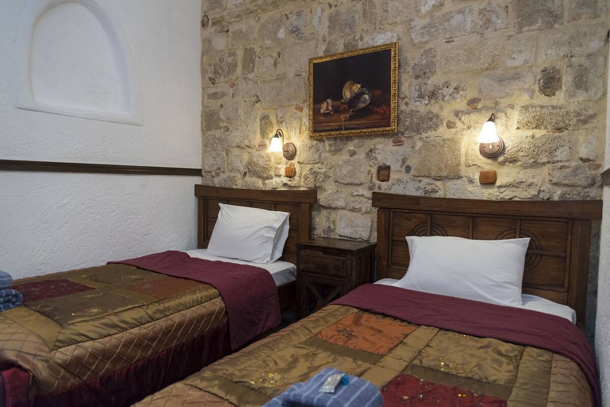Habitación con dos camas, cada una con cabecero de madera y manta roja. Hay luces fijadas en la pared y un cuadro enmarcado sobre las camas.