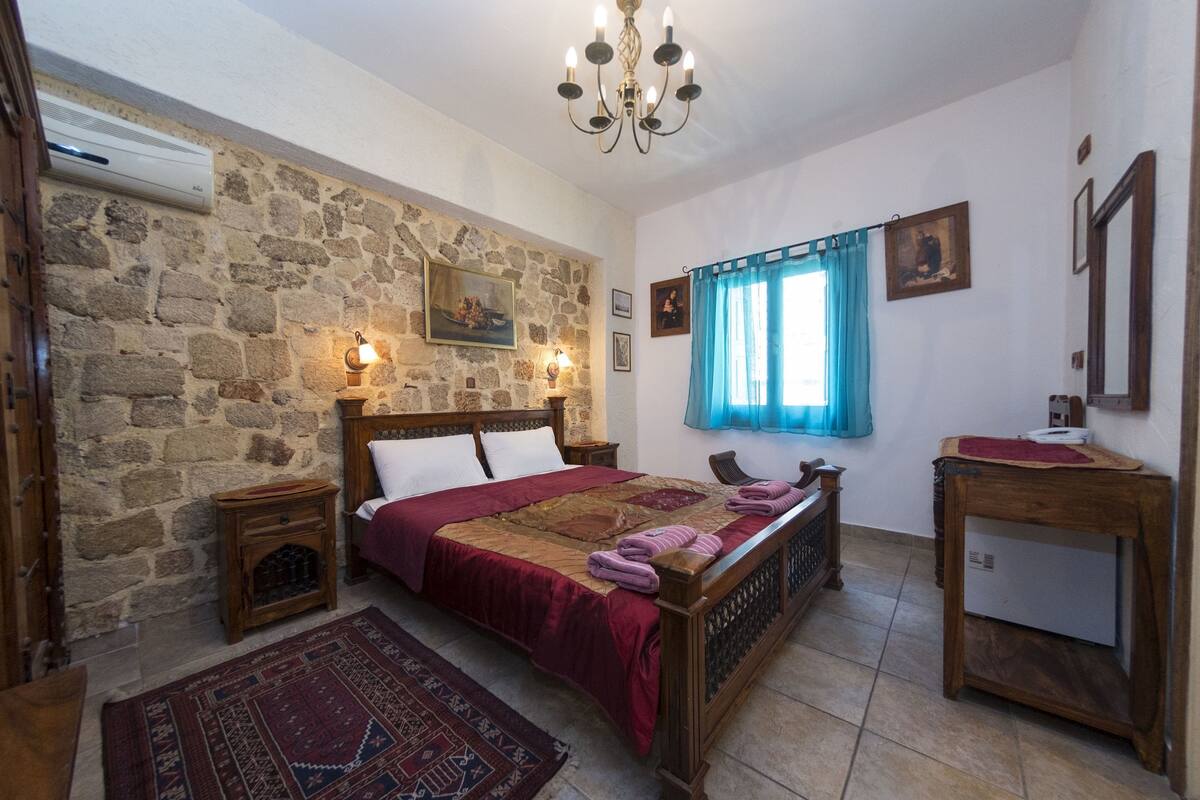 Un dormitorio con pared de piedra, una cama de madera, una mesita de noche, un escritorio y una ventana con cortinas azules.