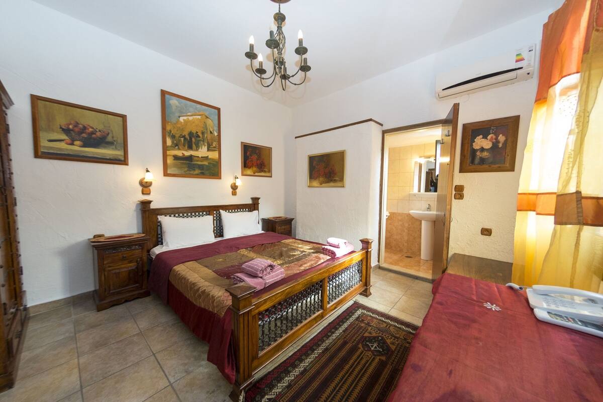 Un dormitorio con cama de madera, una mesita de noche, una lámpara de noche y un baño visible a través de una puerta abierta.