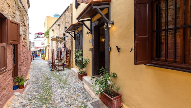 Una stretta strada acciottolata fiancheggiata da edifici, alcuni con piante in vaso e persiane di legno.