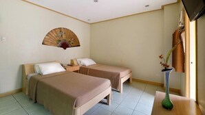 Habitació d'hotel amb dos llits individuals, un ventilador de paret, televisió i un ram de flors sobre una taula de fusta.