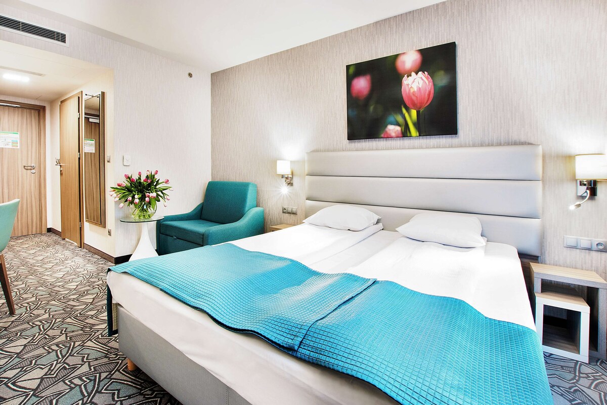 A hotel room with a large bed, a teal armchair, a small table with a vase of flowers, and a painting on the wall.