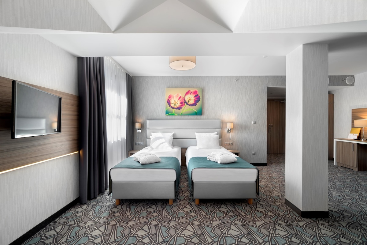 A hotel room with two beds, a large painting above the beds, a flat-screen TV on the wall, and a patterned carpet.