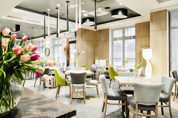 A modern dining area with wooden tables, grey and green chairs, and a vase of pink tulips. - Golden Tulip Krakow Kazimierz (Kraków)