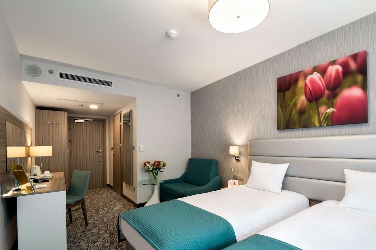 Une chambre d’hôtel avec deux lits, un bureau, une chaise, une petite table ornée d’un vase de fleurs et un tableau représentant des tulipes accroché au mur.