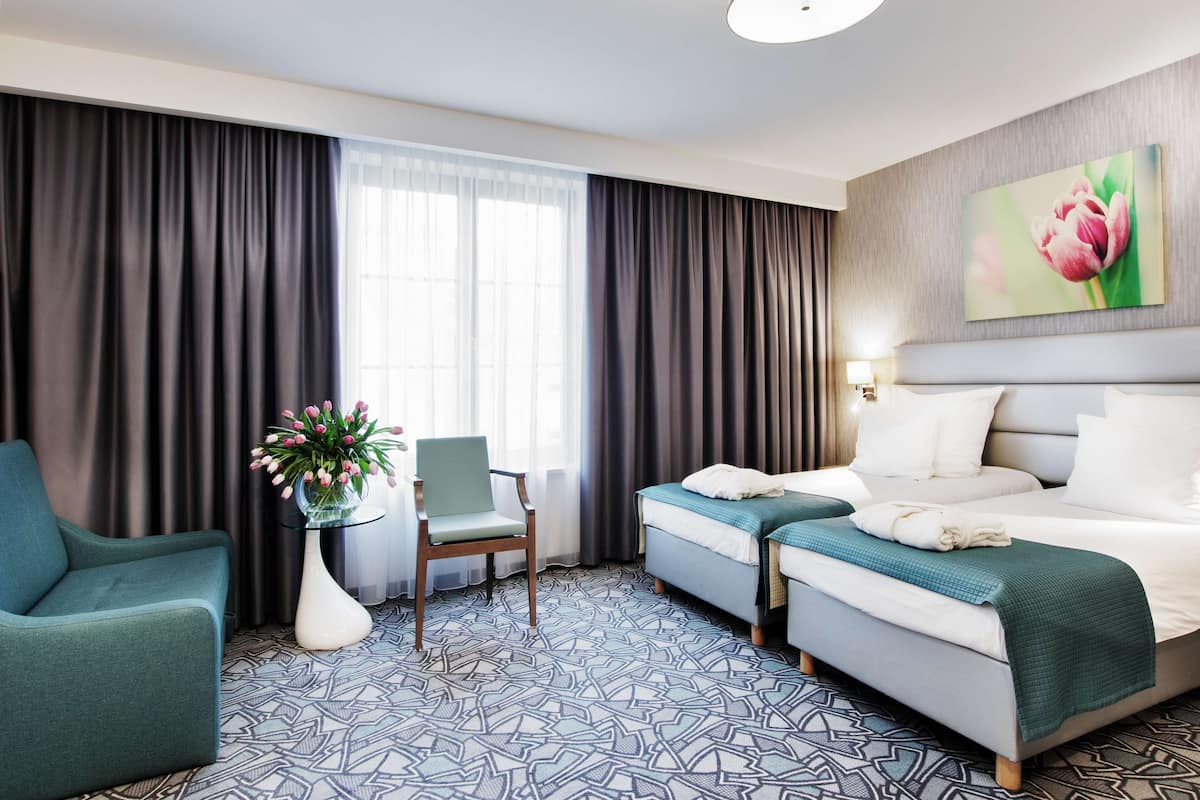 Une chambre d’hôtel avec un grand lit, deux petits lits, un canapé, un fauteuil et un vase de fleurs.