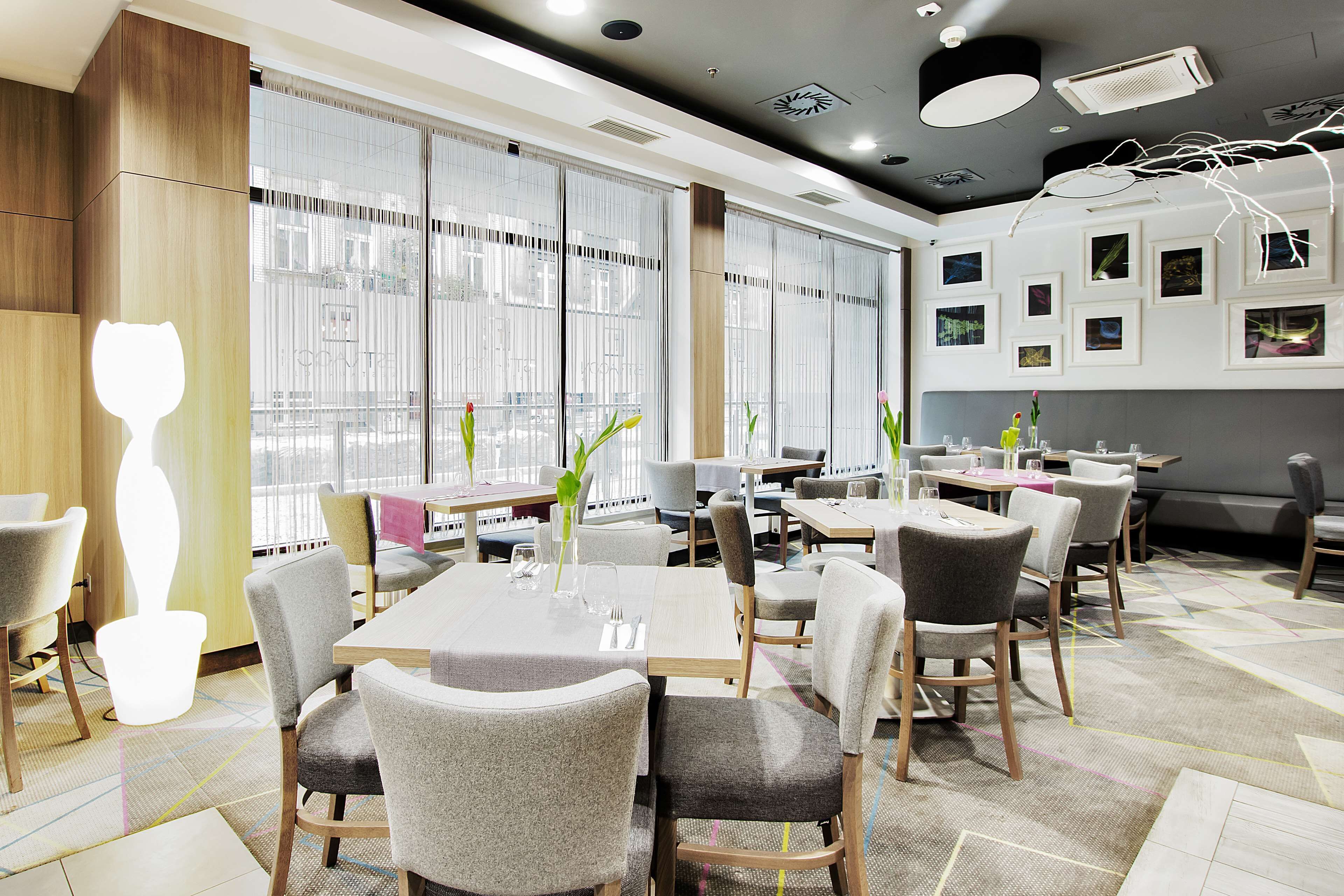 A modern restaurant with multiple tables and chairs, large windows with curtains, and a ceiling with recessed lighting and air conditioning units.