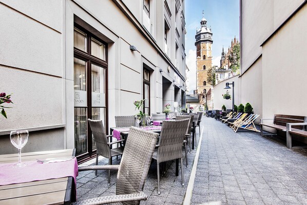 Un espace extérieur aménagé pour les repas, avec des chaises et des tables en osier, un verre à vin, et une vue sur une tour historique en arrière-plan.