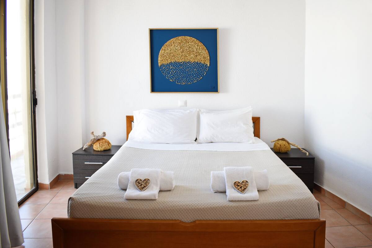 A neatly made bed with white linens and heart-shaped towels, a framed artwork above it, and a wooden headboard.
