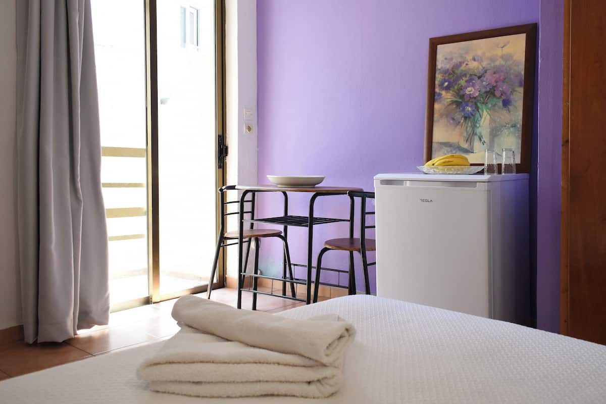 A room with a bed, a small table with a bowl and a banana, a refrigerator, and a painting on the wall.