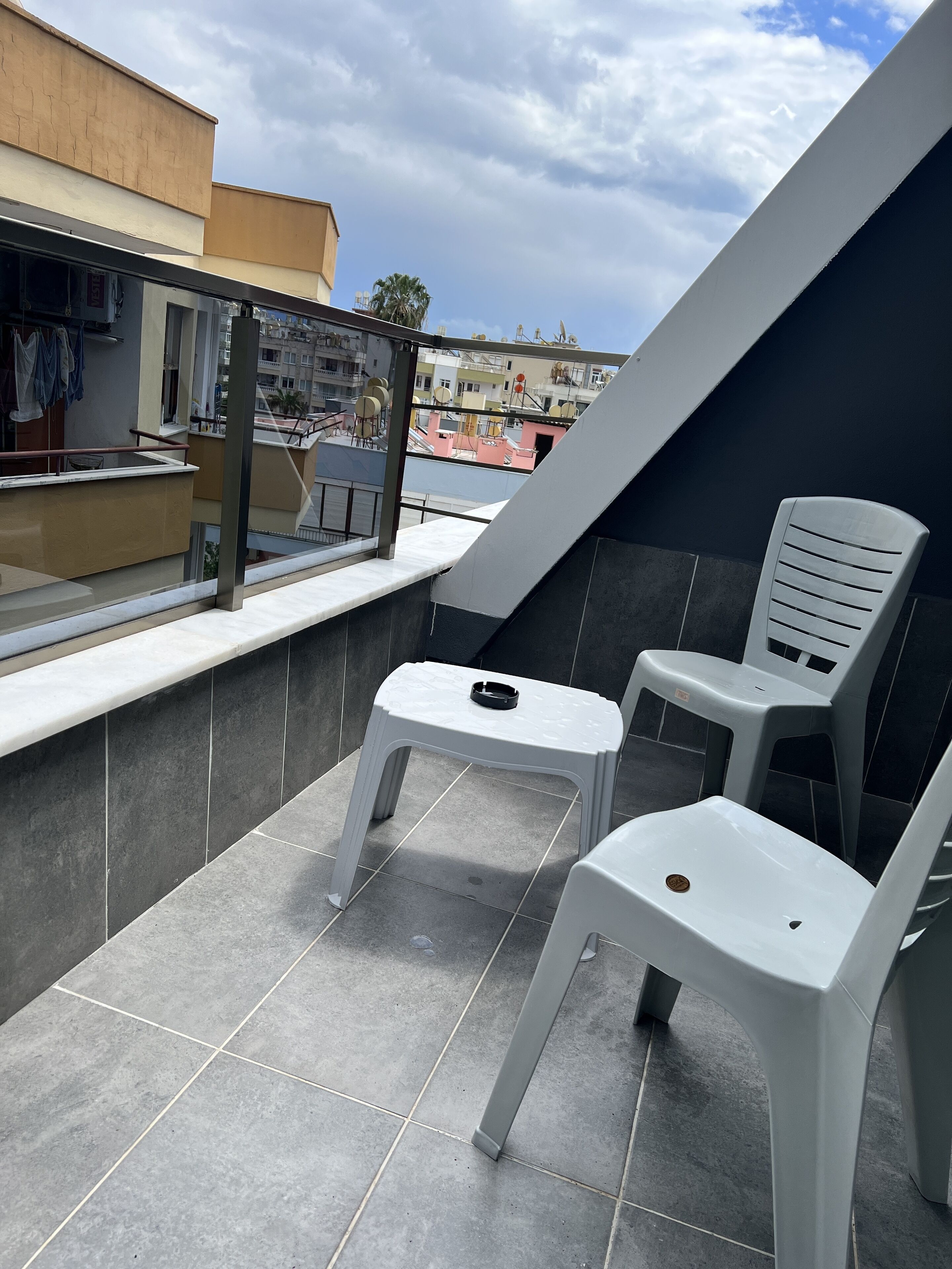 A rooftop terrace with two white plastic chairs, a small table, and a view of the cityscape.