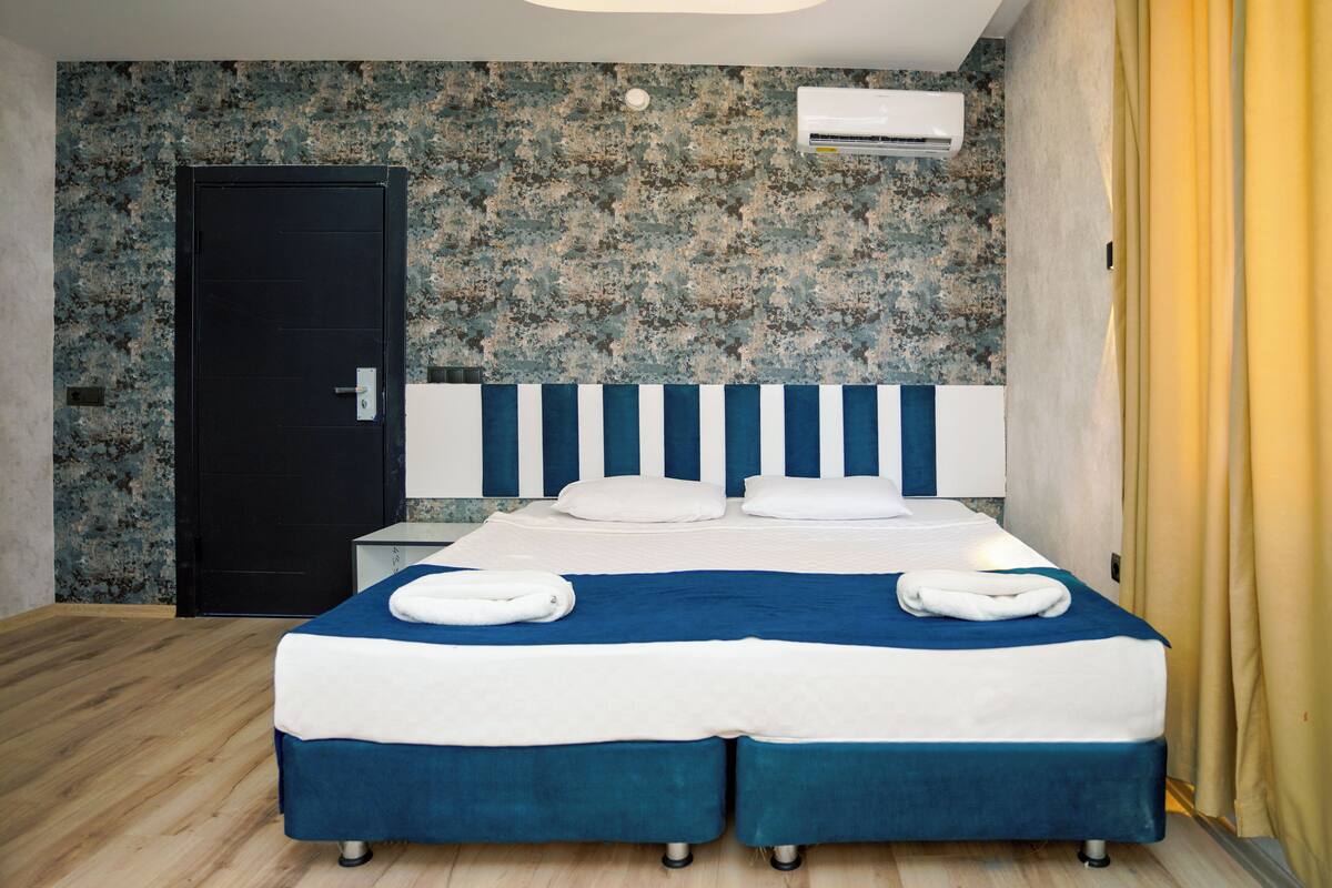 A double bed with a blue and white bedspread, two white pillows, and two towels folded on the bed. There is a black door to the left and an air conditioning unit on the wall above the bed.