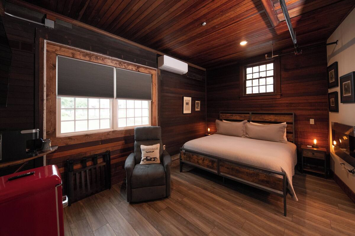 Ein Schlafzimmer mit Holzbalkendecke, einem großen Fenster, einem Bett, einem Sessel und einem roten Koffer.