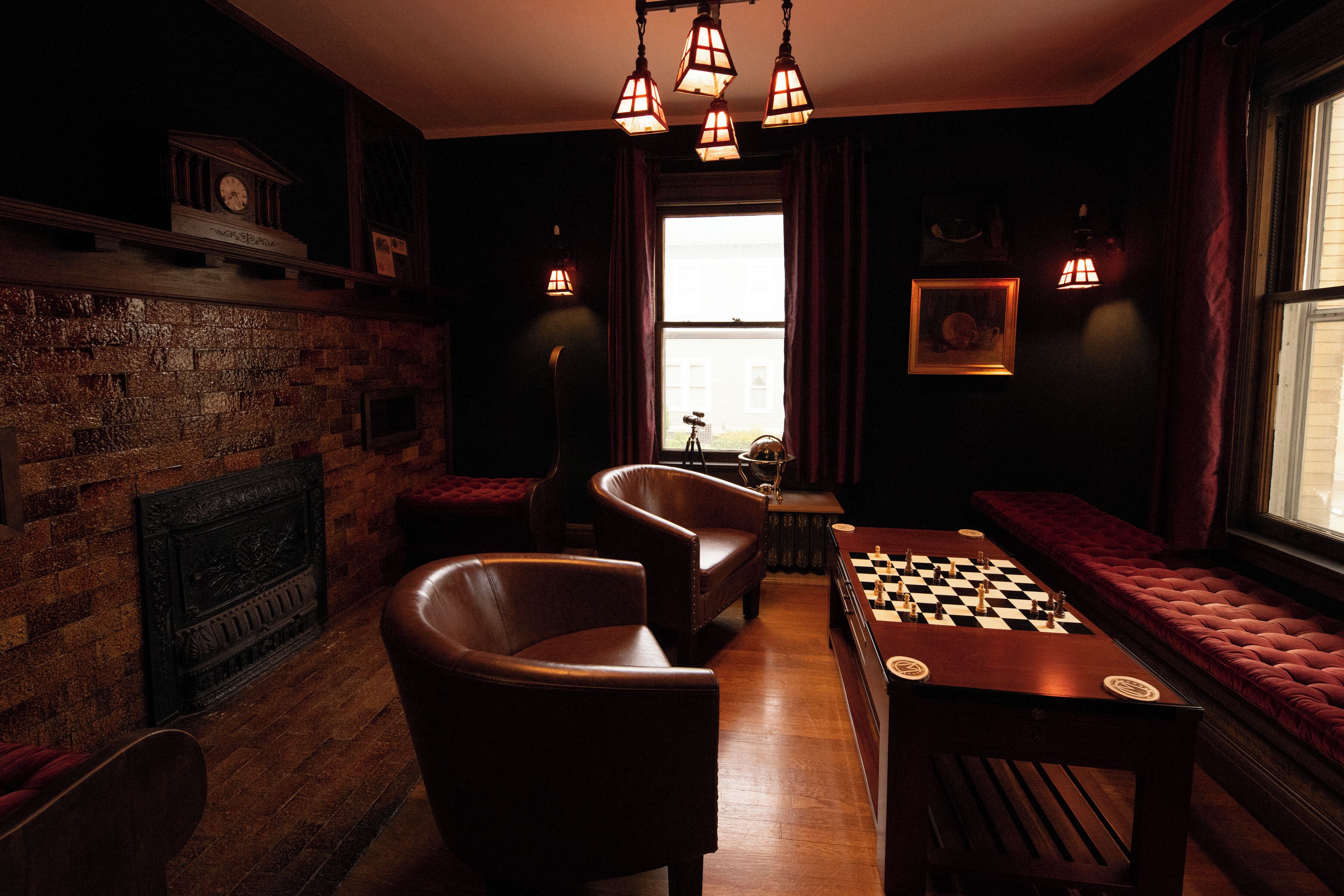 A dimly lit room with a brick fireplace, leather armchairs, a foosball table, and a large window with curtains.