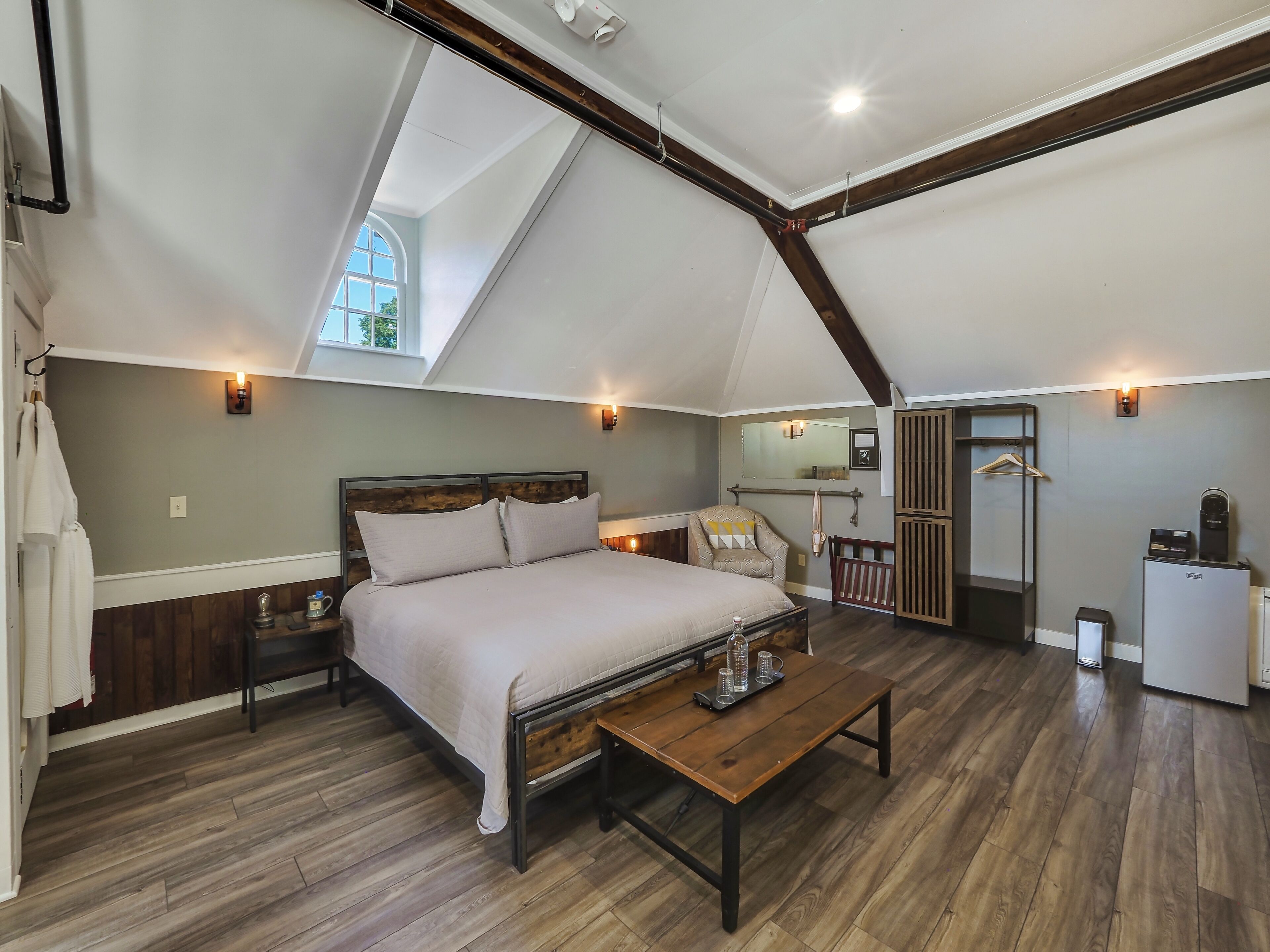A bedroom with a large bed, wooden floor, a small table with glasses, a chair, a wardrobe, and a desk with a computer.
