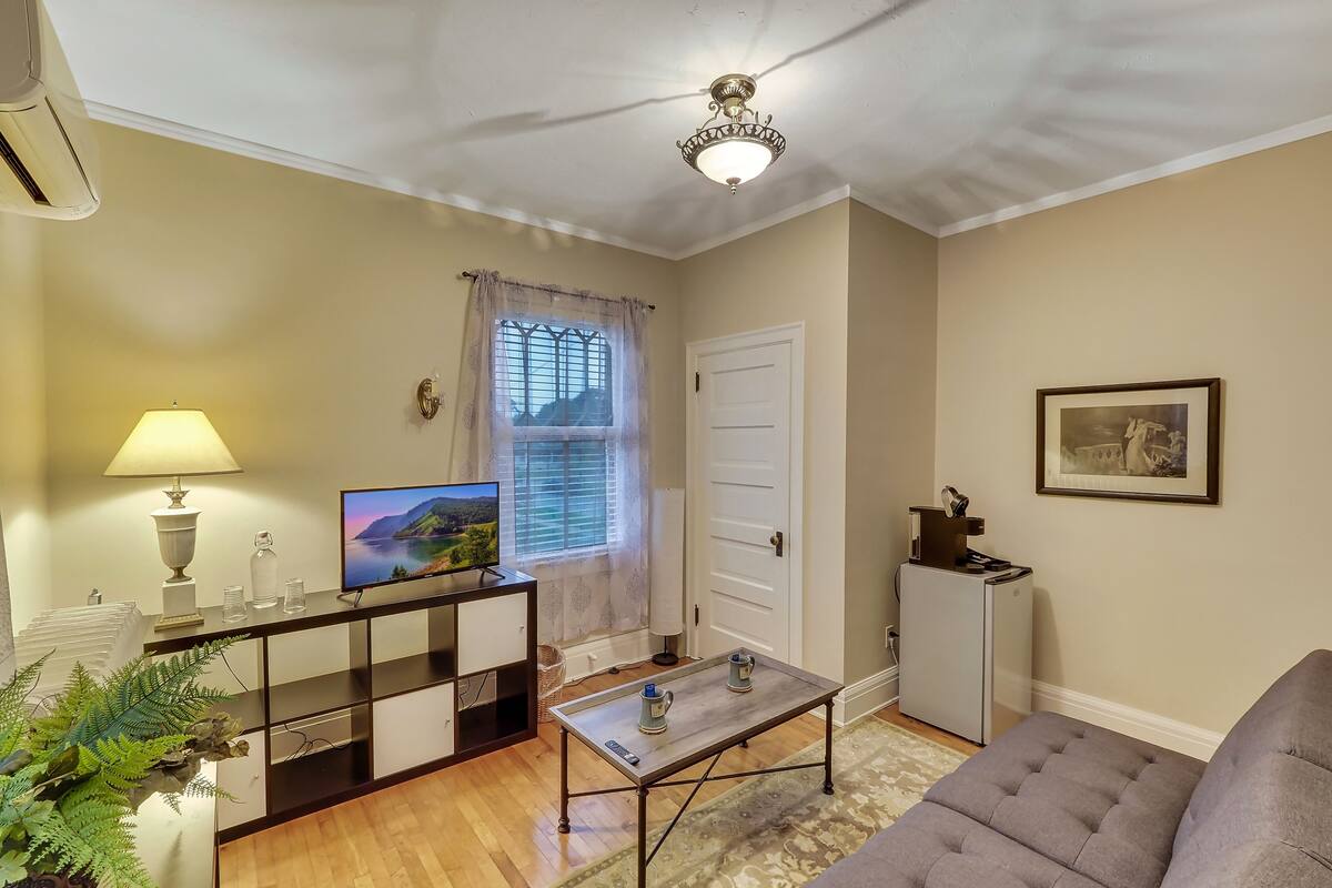 Ein Wohnzimmer mit einer Couch, einem Fernseher, einer Lampe, einem Fenster mit Vorhängen, einer Tür und einem gerahmten Bild an der Wand.