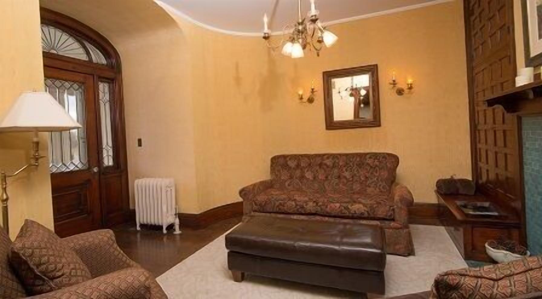 A living room with a sofa, ottoman, and patterned armchairs. There is a radiator, a chandelier, and a mirror on the wall.