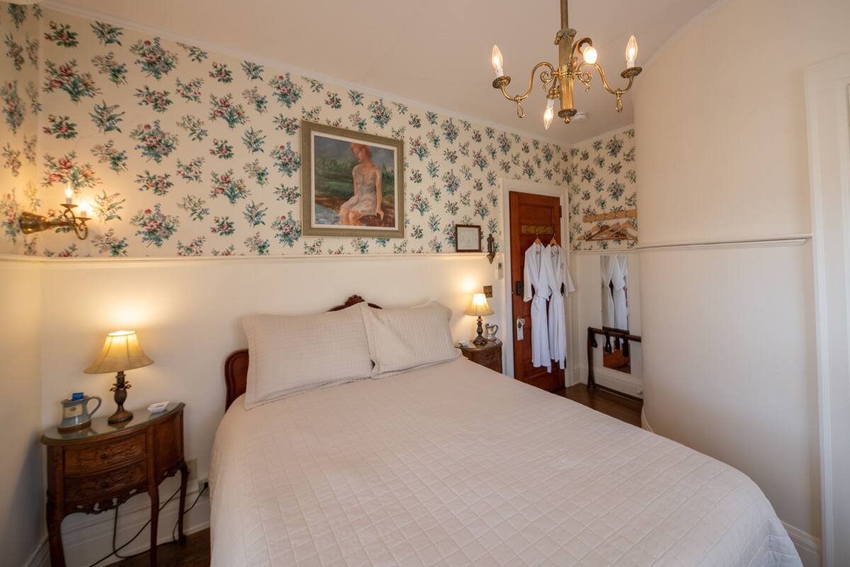 Ein Schlafzimmer mit floralem Wandpapier, einem Bett mit weißer Bettwäsche, einer Kristallleuchtre, einem Nachttisch mit Lampe und einem Kleiderständer mit Kleidung.