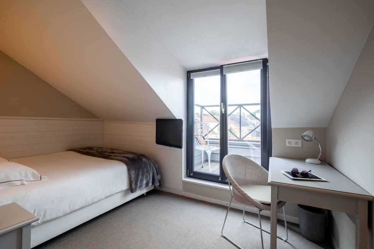 A small, modern bedroom with a bed, a desk, a chair, a TV, and a window with a view of buildings.