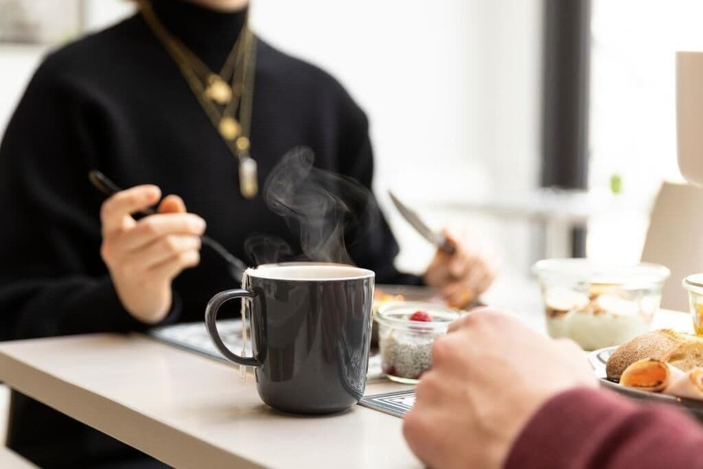Una persona sentada a una mesa con una taza de café, un plato de comida y un recipiente de vidrio.