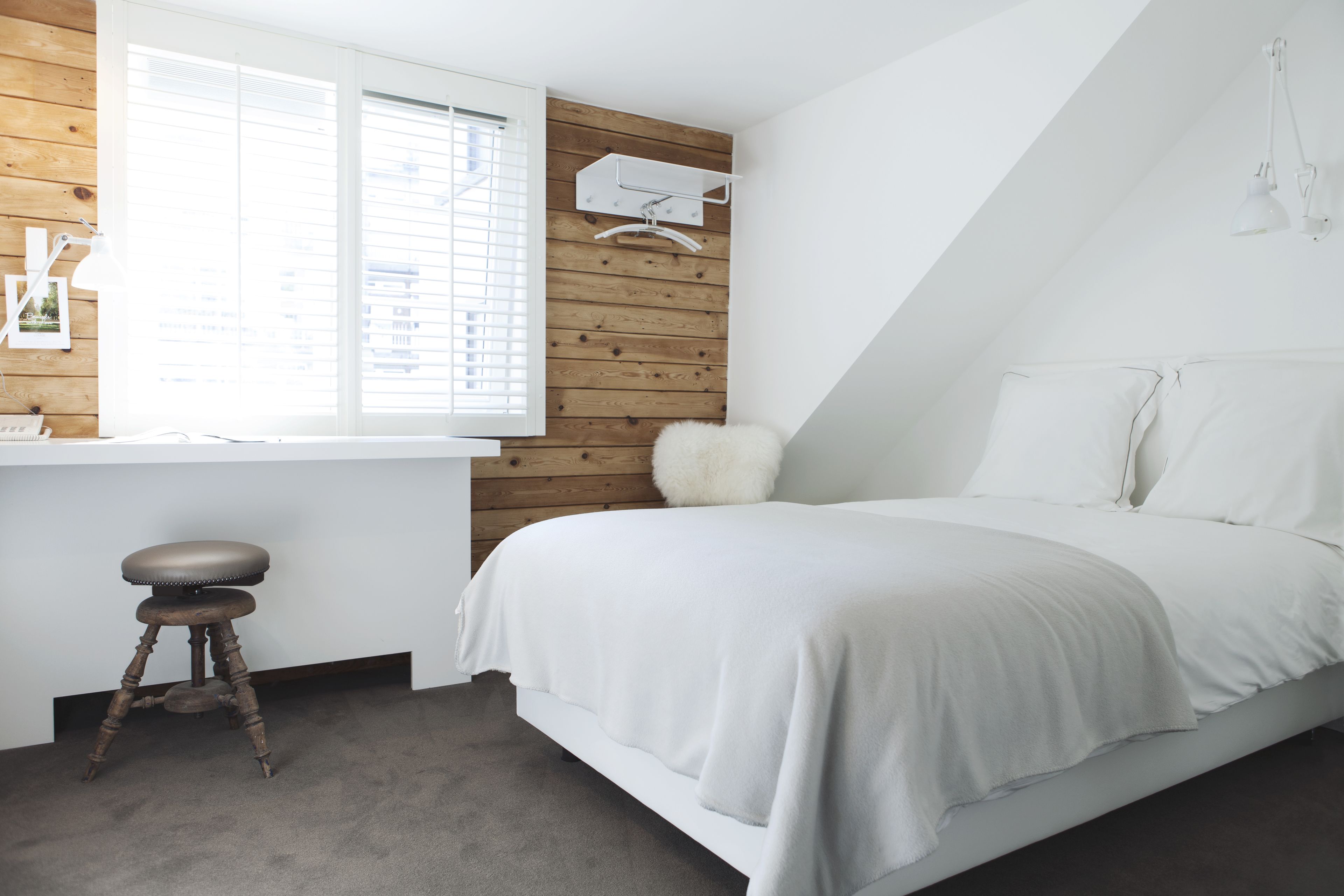 A bedroom with a bed, a desk with a chair, a window with blinds, a hanging light fixture, and a wooden wall panel.