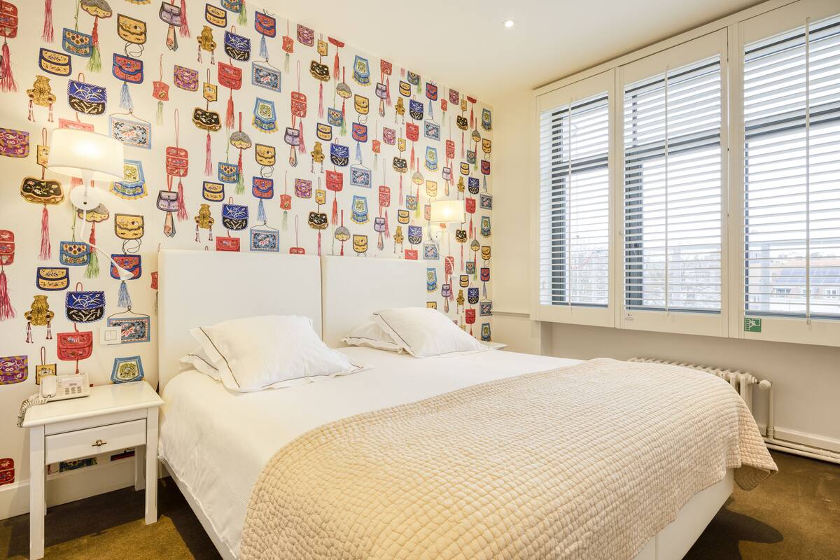 A bedroom with a bed, white nightstands, a wall decorated with various colorful items, and a window with blinds.