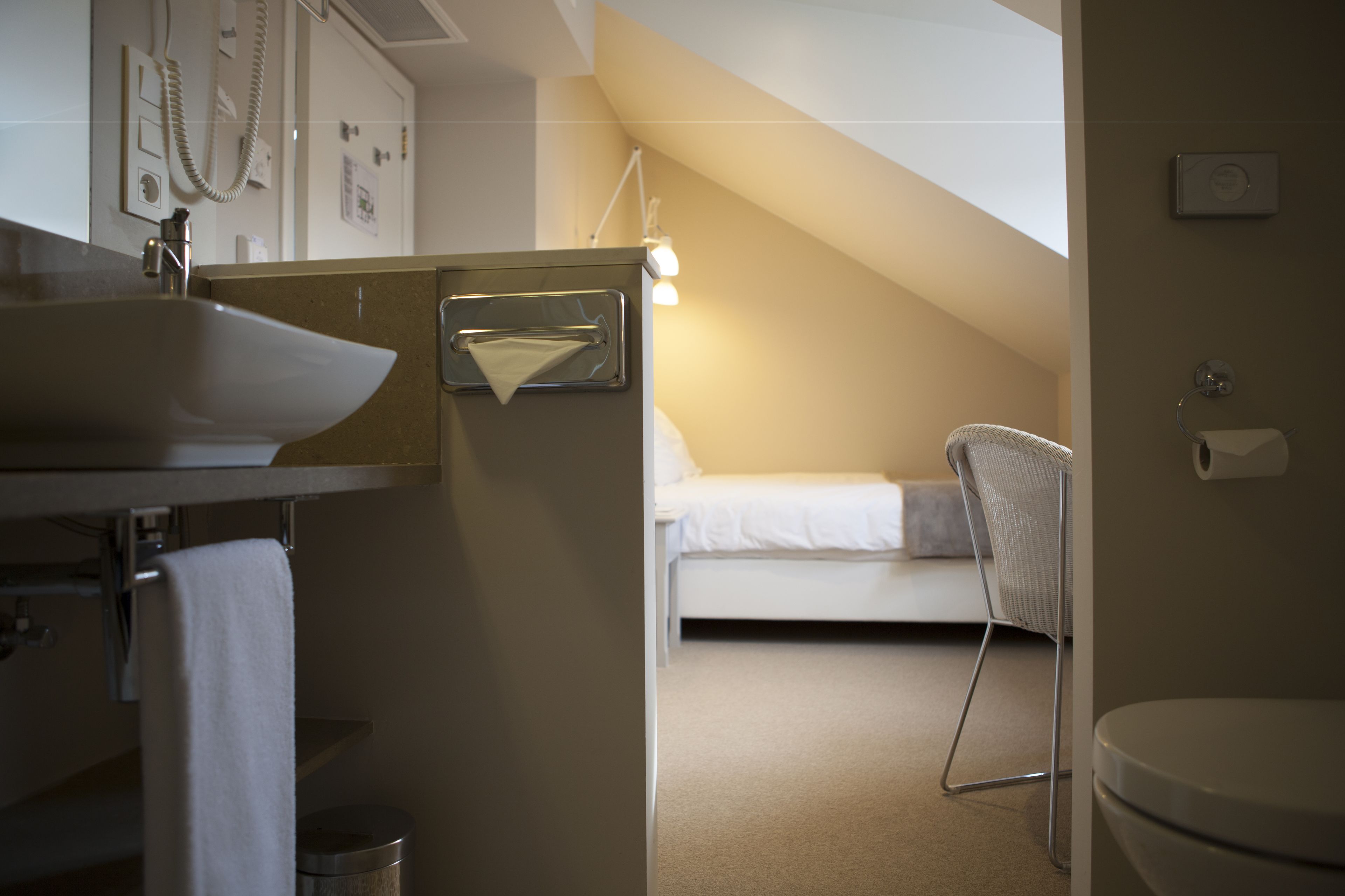 Une salle de bain moderne et compacte, équipée d’un lavabo, d’un porte-serviettes et de toilettes. On aperçoit un lit à l’arrière-plan, ce qui laisse supposer qu’il s’agit d’une petite pièce, peut-être partagée.