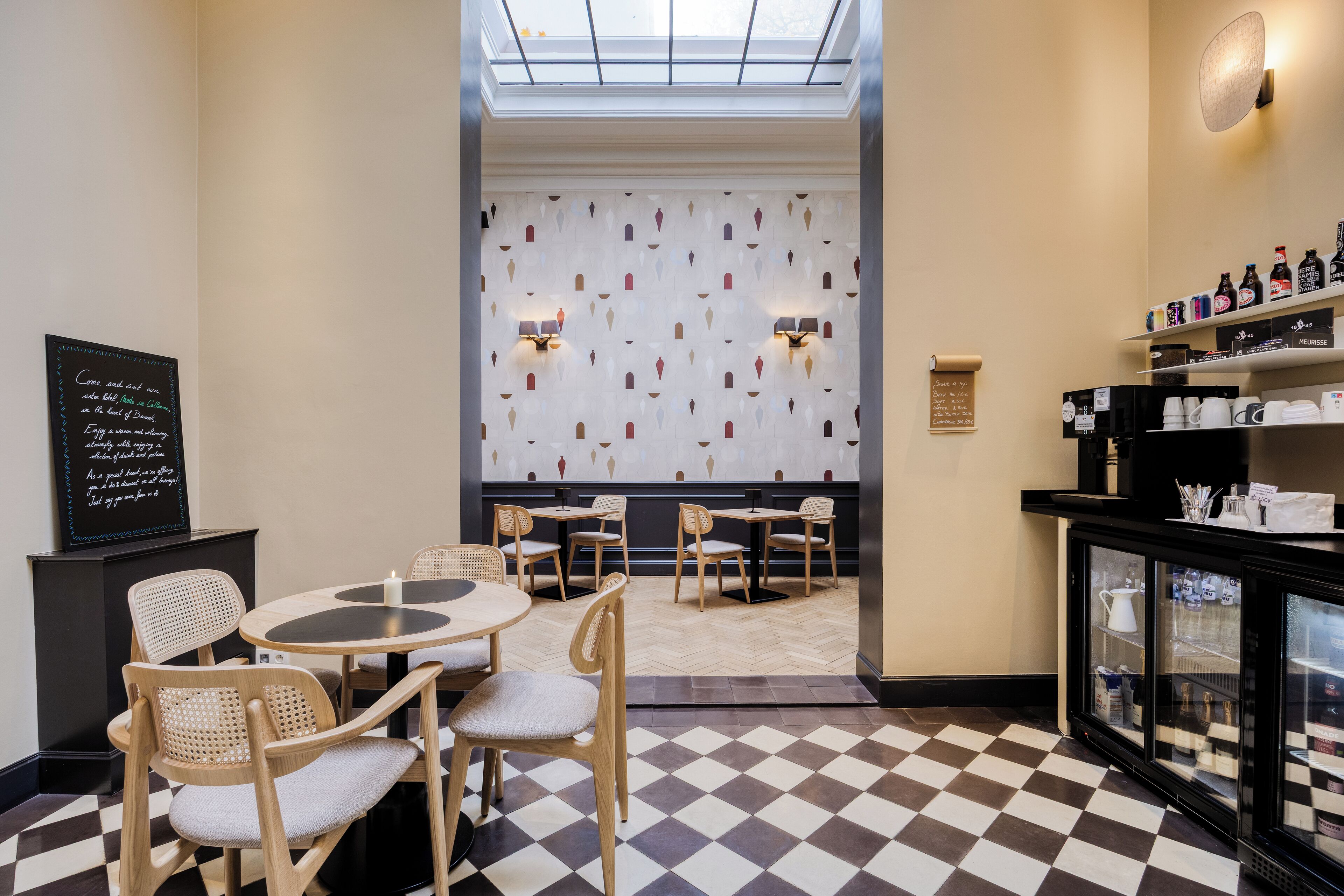 A restaurant with a checkered floor, wooden chairs, and tables. There is a wall with a pattern and two wall-mounted lights.