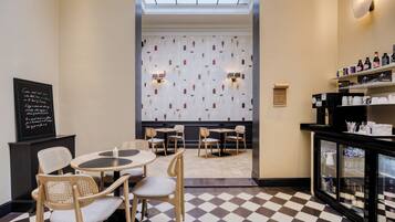 A restaurant with a checkered floor, wooden chairs, and tables. There is a wall with a pattern and two wall-mounted lights.