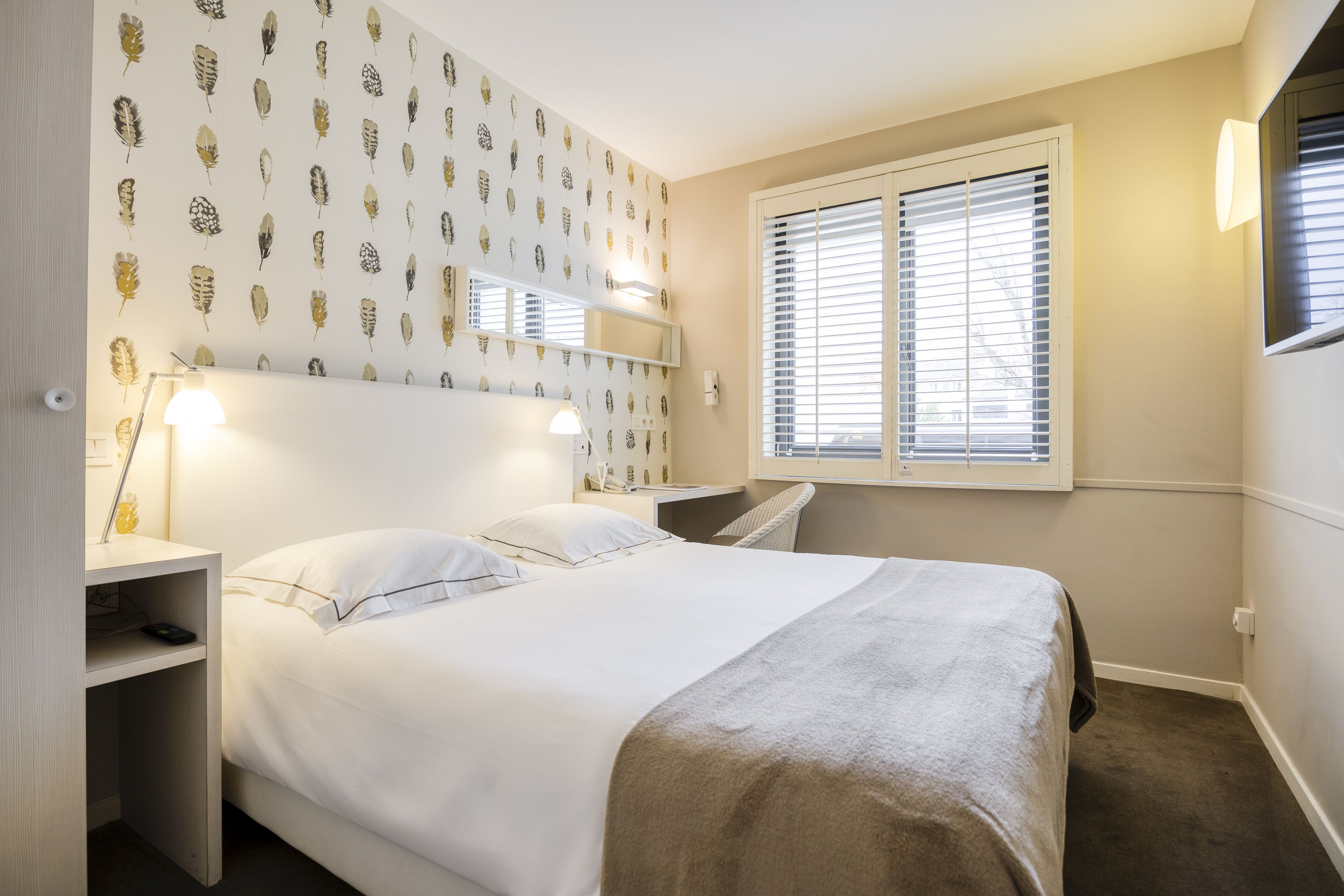 Una habitación de hotel ordenada con una cama, mesitas de noche, un escritorio y una ventana con persianas.