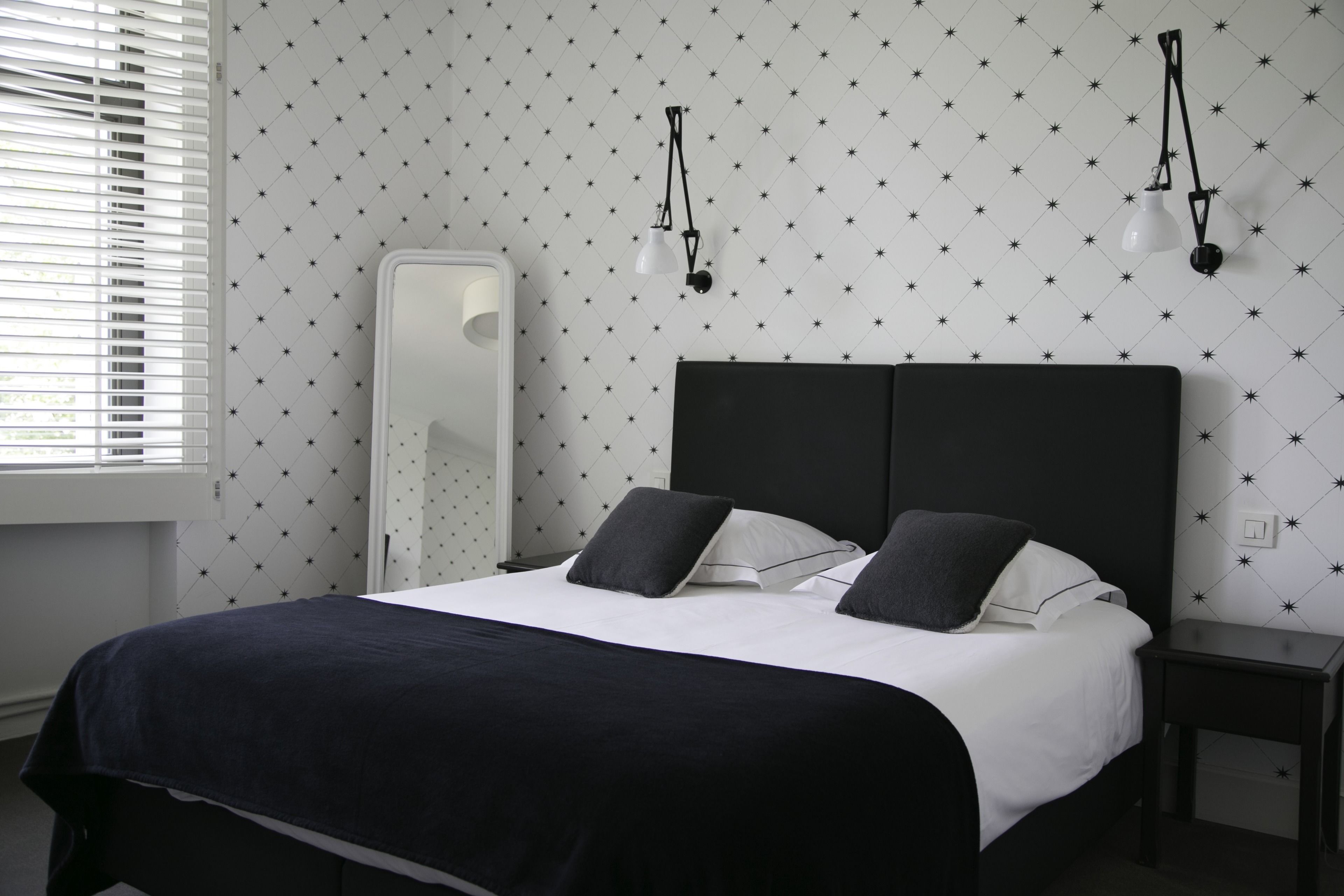 A neatly made bed with a black headboard and white bedding, a bedside table, a wall-mounted lamp, and a window with blinds.