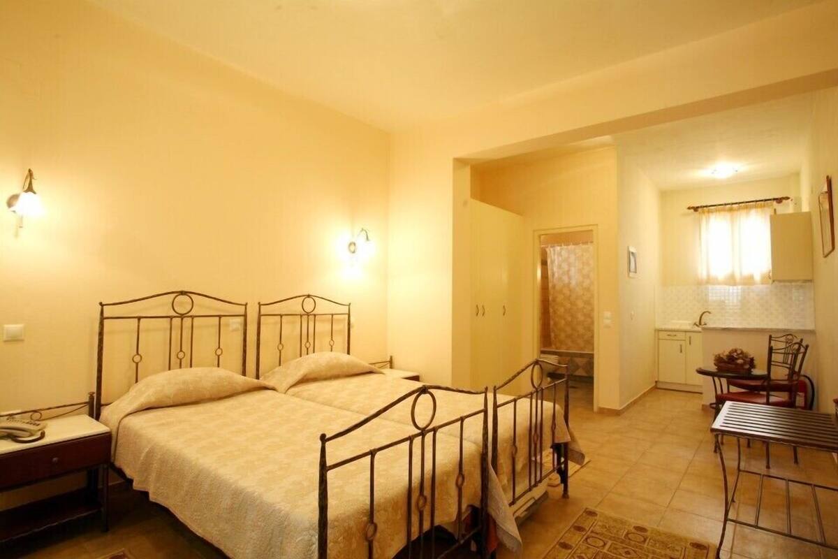 A bedroom with a metal bed frame, a bedside table, and a small desk with a chair. There is a kitchenette with a sink and a mirror.