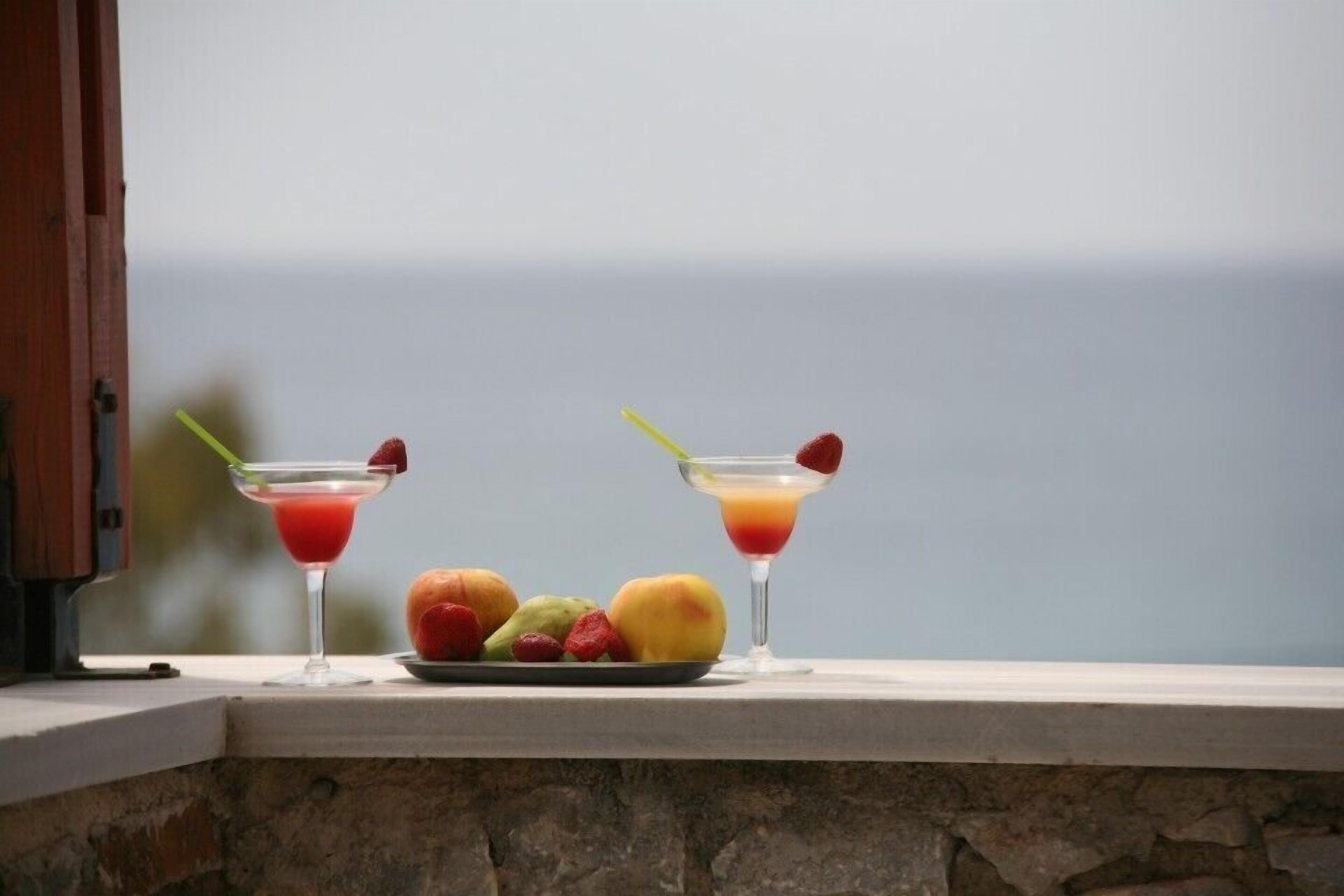 Dos copas de martini con adornos de frutas y un plato de frutas variadas sobre un borde de piedra.