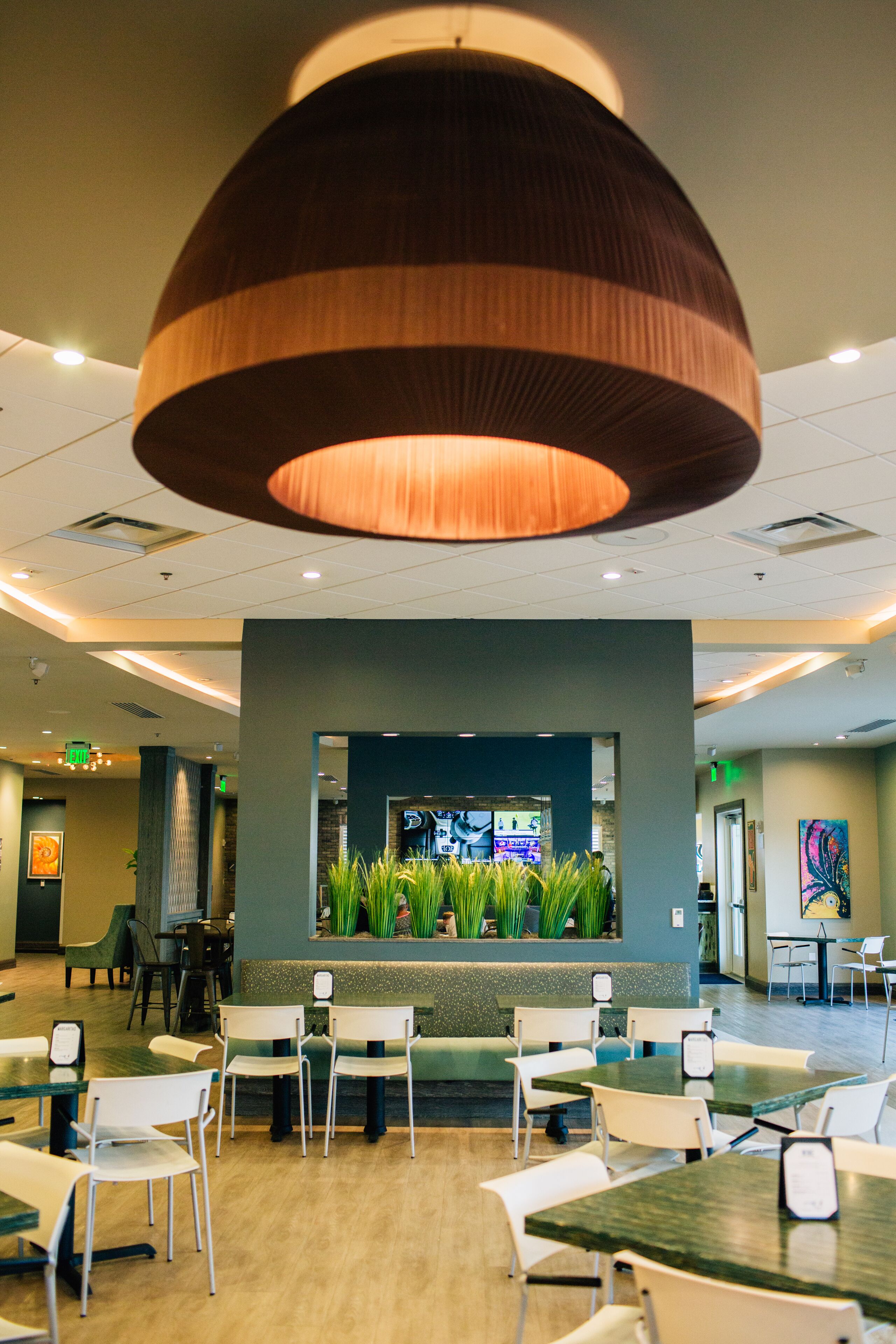 A modern restaurant interior with a central seating area, a large hanging light fixture, and a wall-mounted screen displaying content.