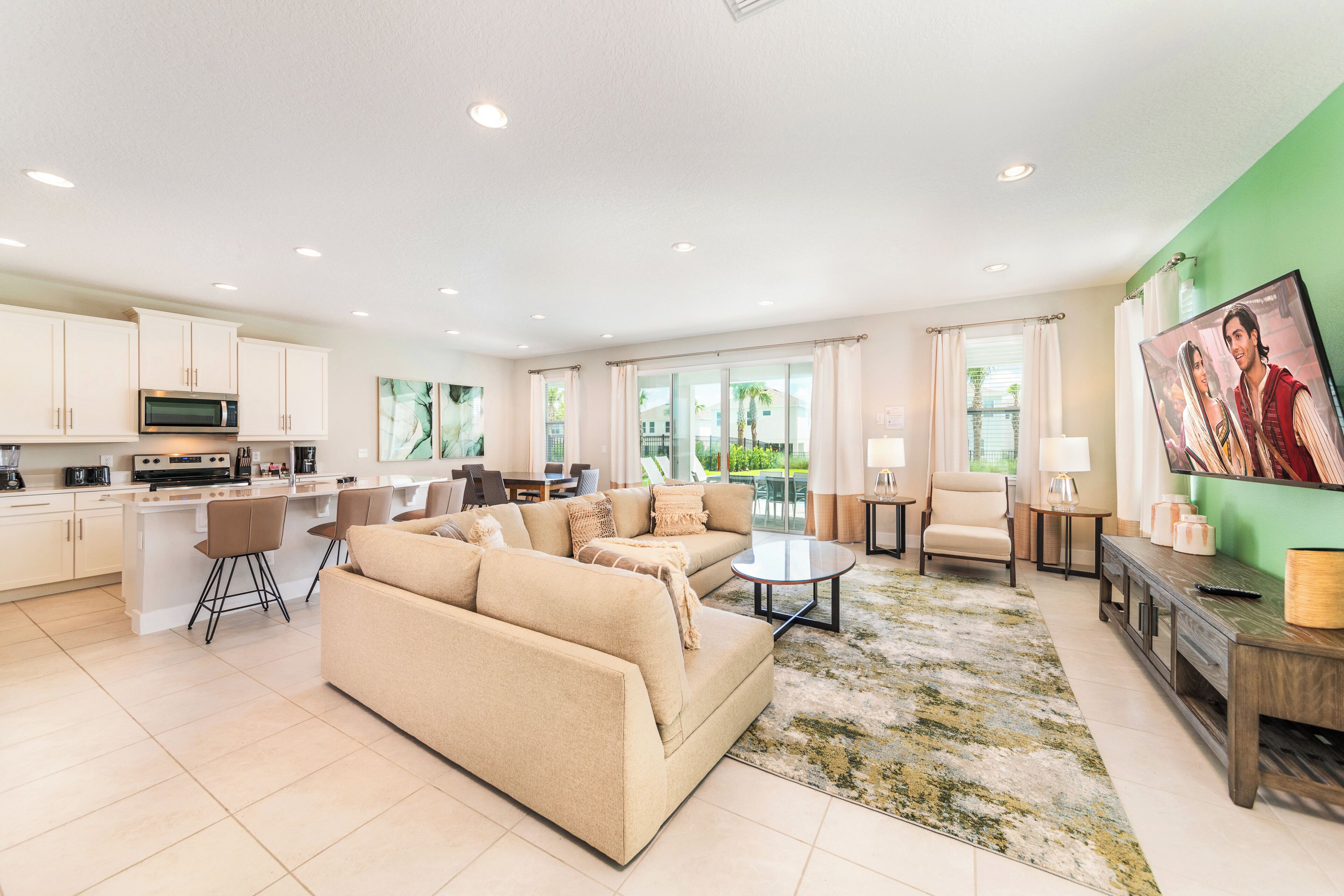 A modern living room with a beige sofa, a coffee table, and a TV. There is a kitchen area with white cabinets and a microwave.