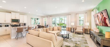 A modern living room with a beige sofa, a coffee table, and a TV. There is a kitchen area with white cabinets and a microwave.