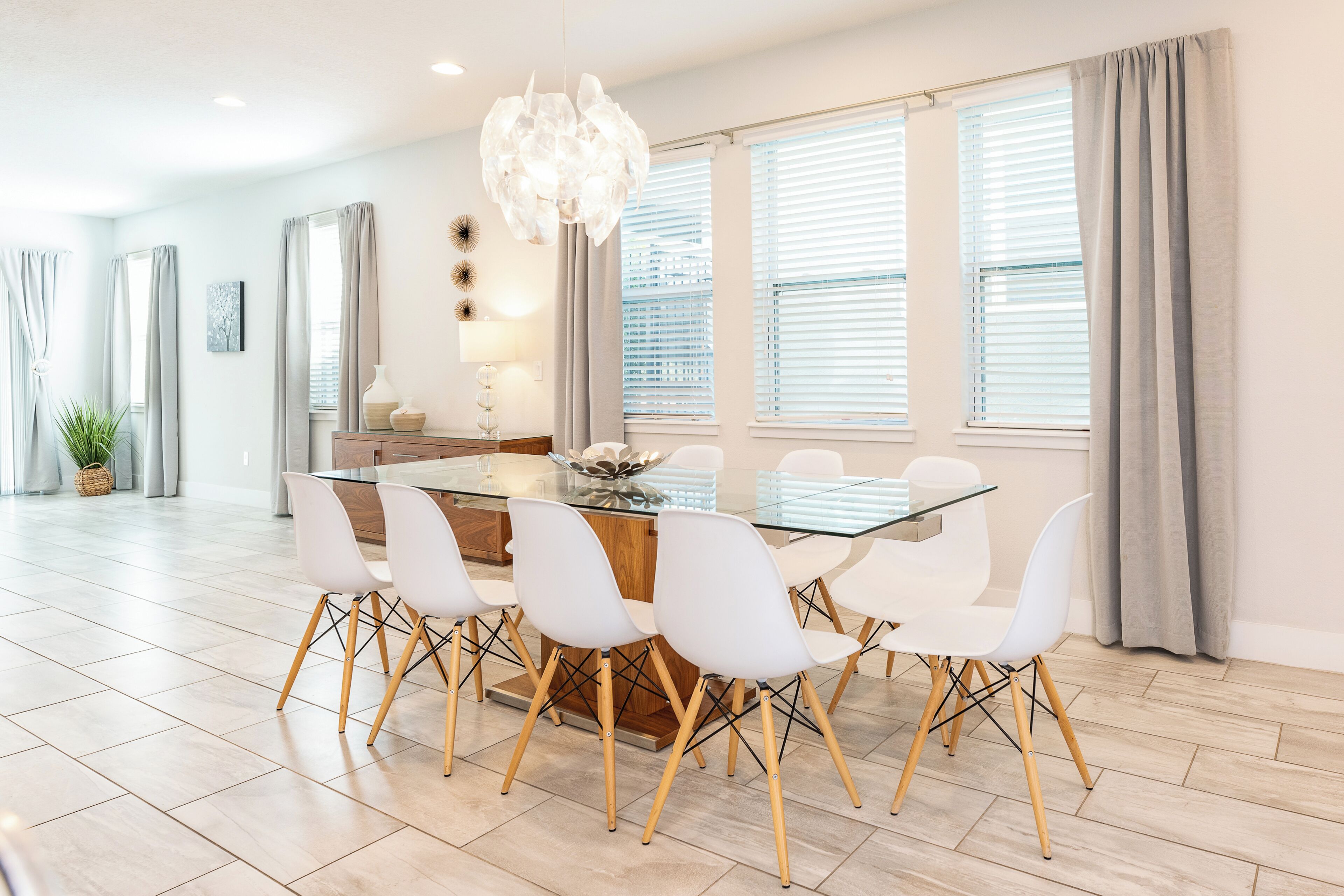 A modern dining area with a glass table and white chairs, large windows with blinds, and a decorative chandelier.