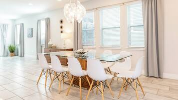 A modern dining area with a glass table and white chairs, large windows with blinds, and a decorative chandelier.