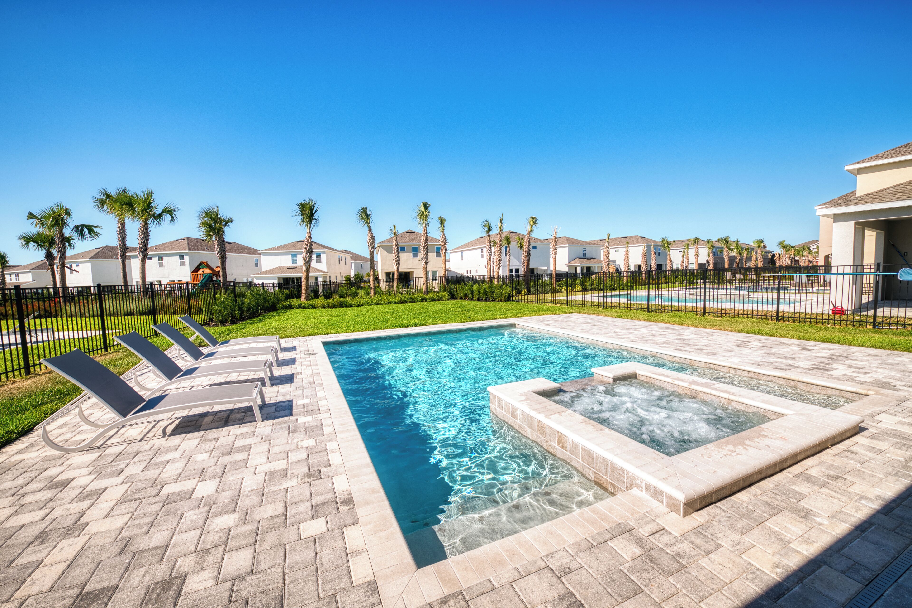 A swimming pool with lounge chairs, a hot tub, and a fenced area with palm trees.