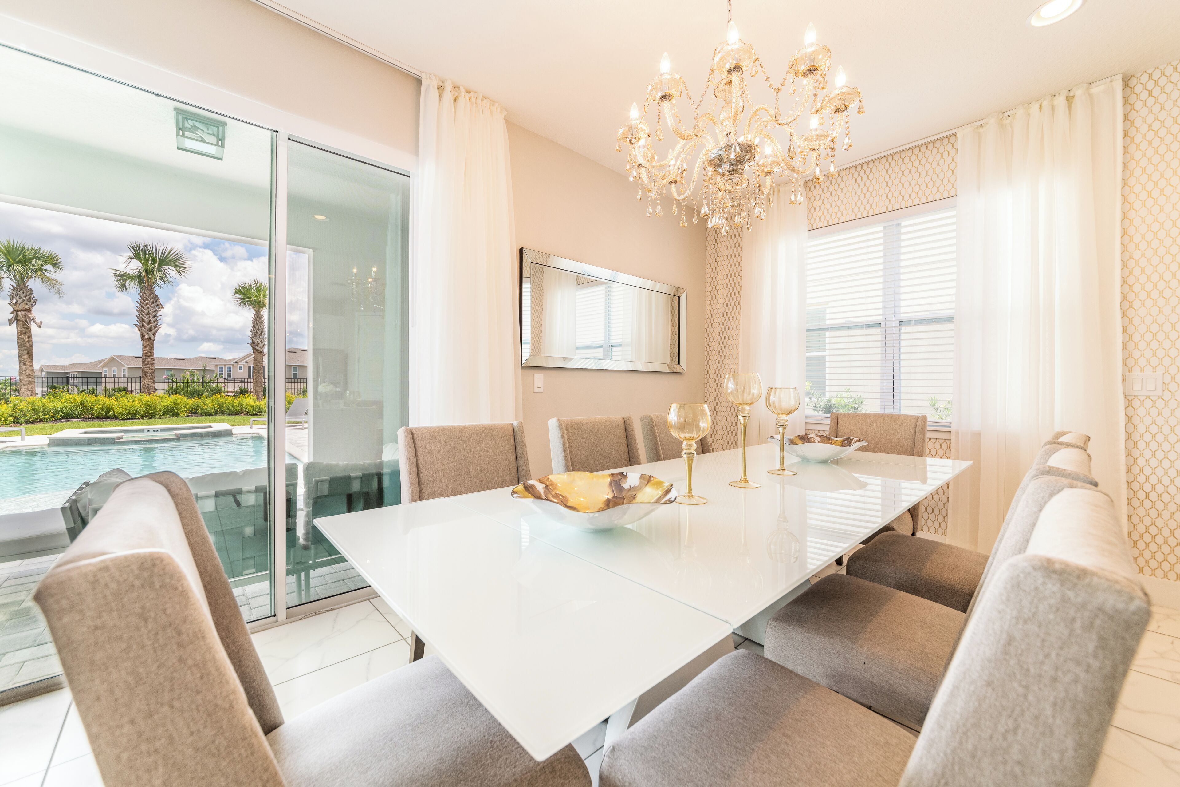 A modern dining room with a glass table, beige chairs, a chandelier, and a view of a swimming pool.