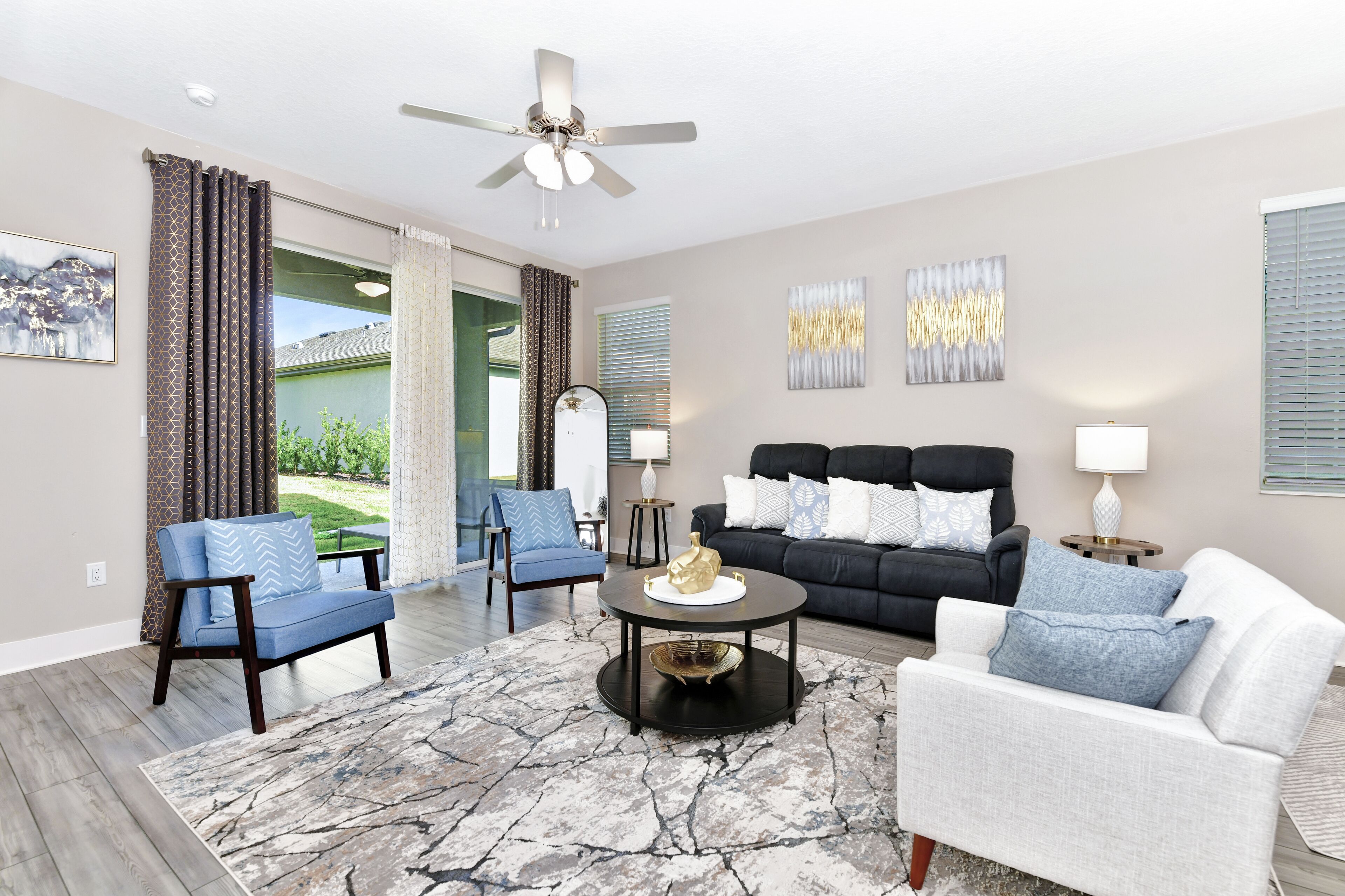 Un salón con ventilador de techo, un sofá negro, un sofá blanco y un sillón azul. Hay una mesa de centro con un cuenco decorativo y una lámpara. La habitación tiene un ventanal con cortinas.