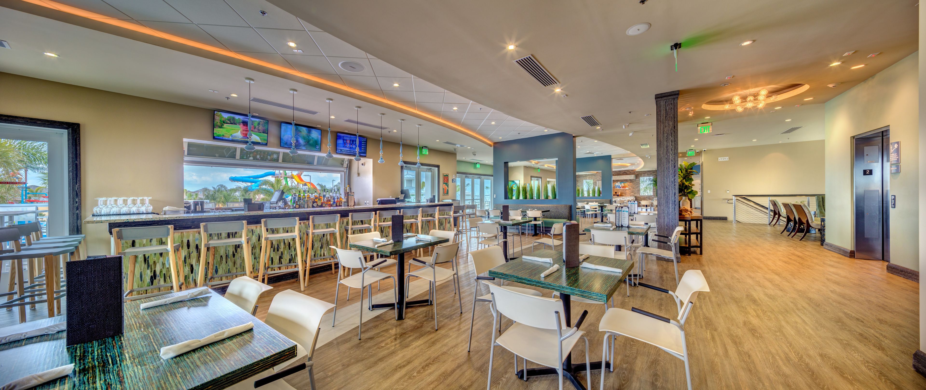 A modern restaurant with multiple seating areas, a bar, and large windows offering views of the outdoor area.