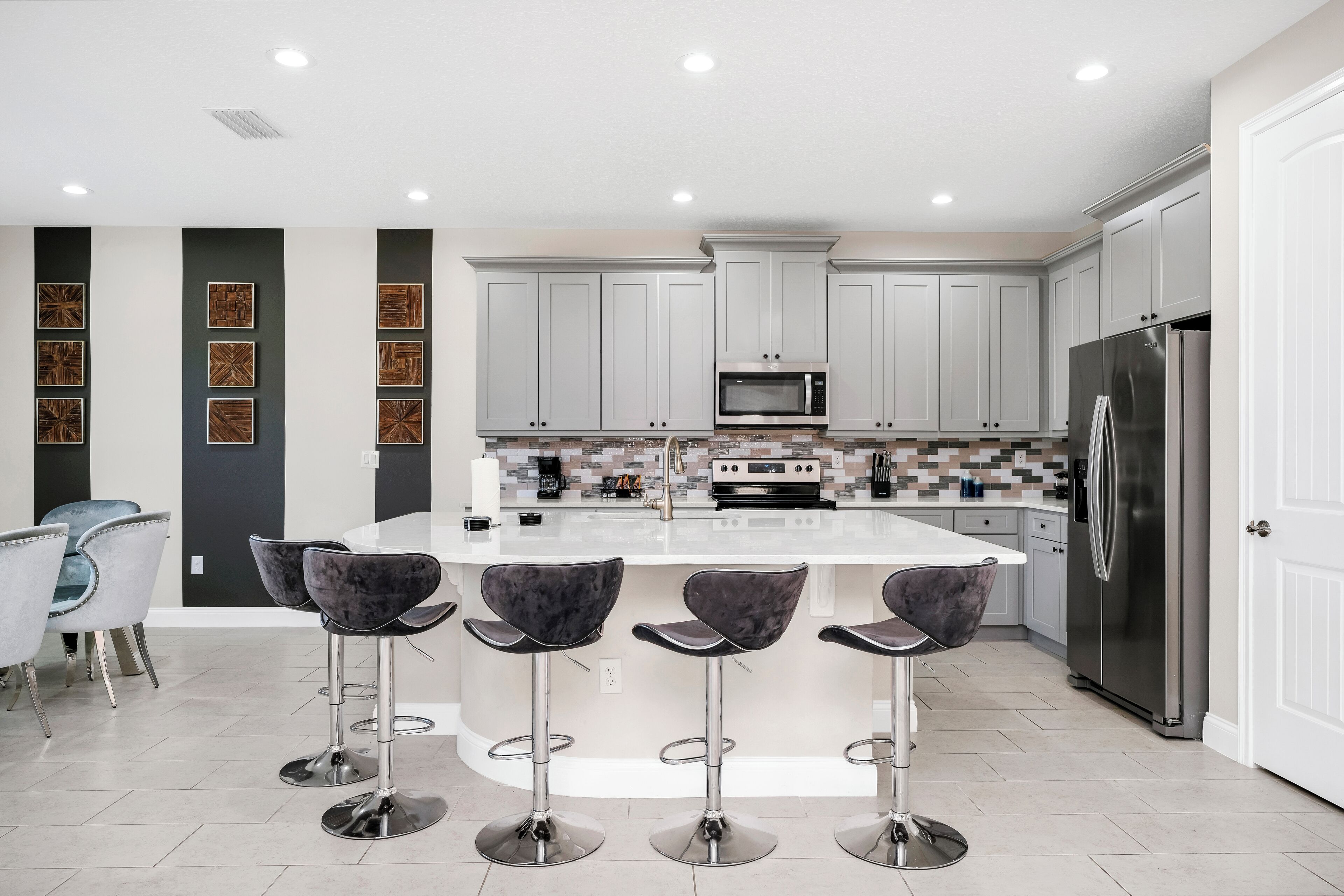 A modern kitchen with grey cabinets, a stainless steel refrigerator, a microwave, and a central island with bar stools.
