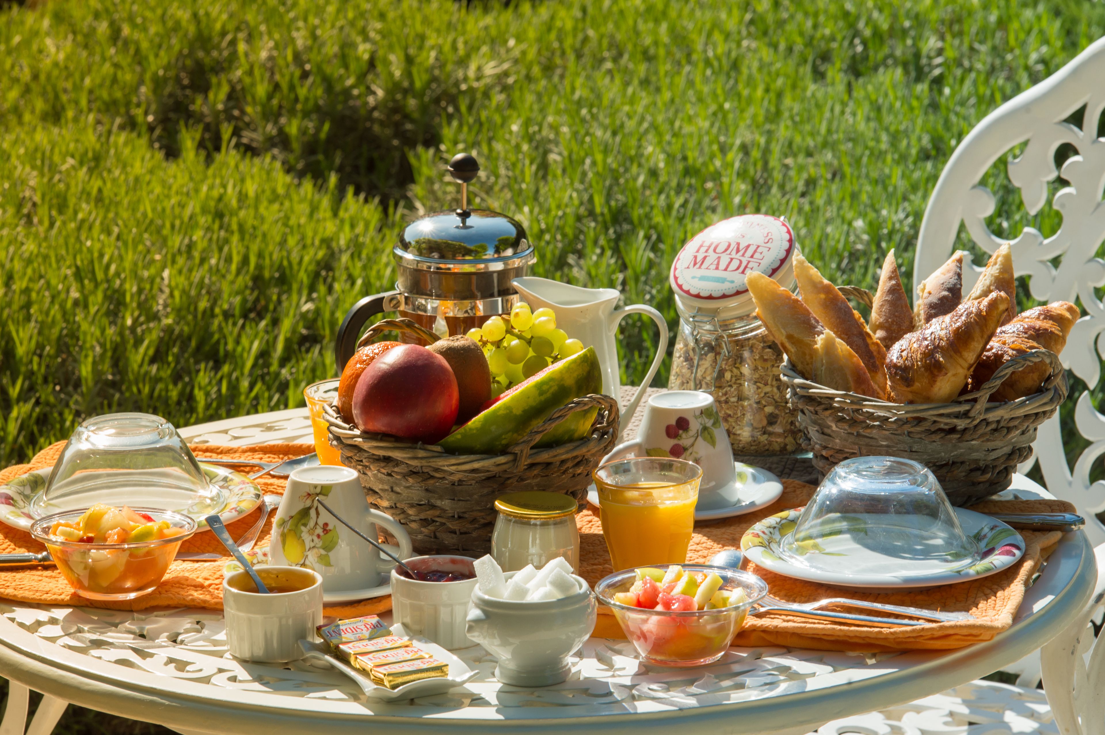 Un desayuno preparado con cafetera, frutero, jugo y pan sobre una mesa al aire libre.