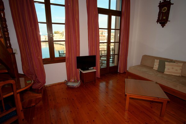 Un salón con suelo de madera, un sofá, un televisor sobre un mueble bajo y ventanas grandes con cortinas.